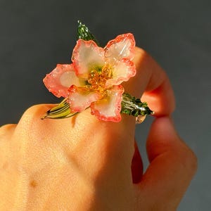 May include: A floral ring with a flower design. The ring has pink and white petals, with gold accents. Green leaves and stems complete the natural look. A unique piece of jewellery.