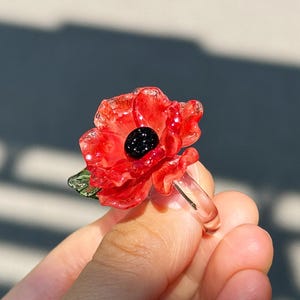 May include: A clear ring with a red poppy flower, black centre, and green leaf detail. The ring is held up, displaying the intricate floral design. A unique piece of jewellery.