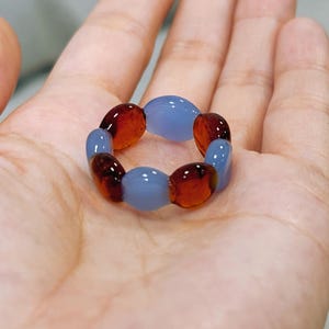 Könnte beinhalten: Ein Ring aus abwechselnd oval geformten Perlen. Der Ring hat durchscheinende, hellblaue und bernsteinfarbene Perlen. Der Ring wird auf der Handfläche einer Person gezeigt.