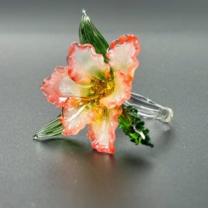 May include: A delicate glass flower sculpture with a clear stem and green leaves. The flower has white petals with red edges and a yellow center. The glass art piece is on a gray surface.
