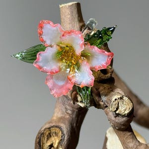 May include: A handcrafted glass flower with white petals edged in pink, a yellow centre, and green leaves. The flower is mounted on a piece of natural wood, creating a unique decorative piece.