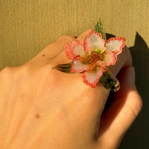 May include: A flower ring with a translucent design. The ring features a white and pink flower with green leaves. The ring is worn on a hand, with a shadow in the background.