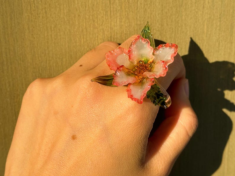 May include: A flower ring with a translucent design. The ring features a white and pink flower with green leaves. The ring is worn on a hand, with a shadow in the background.