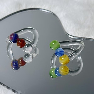 May include: Two clear glass rings, each with a series of colorful glass beads. One ring features red, blue, and clear beads, while the other has green, blue, and yellow beads. The rings are displayed on a reflective surface.