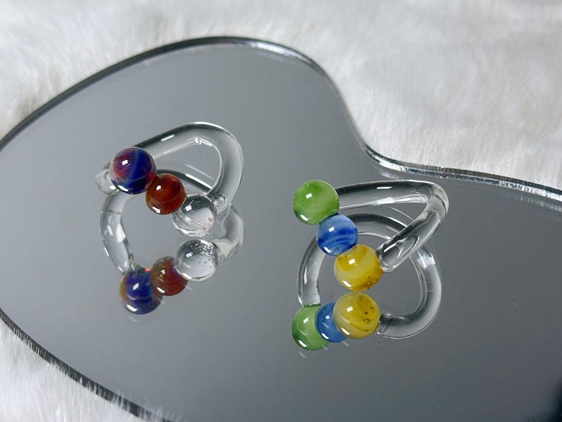 May include: Two clear glass rings, each with a series of colorful glass beads. One ring features red, blue, and clear beads, while the other has green, blue, and yellow beads. The rings are displayed on a reflective surface.