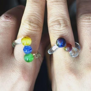 May include: Two clear glass rings, each adorned with colorful glass beads. One ring features yellow, blue, and green beads, while the other has blue, red, and clear beads. The rings are designed with a unique, artistic flair.