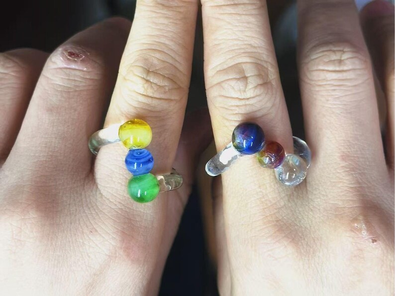May include: Two clear glass rings, each adorned with colorful glass beads. One ring features yellow, blue, and green beads, while the other has blue, red, and clear beads. The rings are designed with a unique, artistic flair.