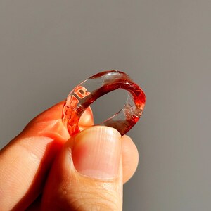 Pode incluir: Um anel de resina transparente com detalhes vermelhos e a palavra "ae" incorporada no design. O anel &eacute; exibido em um fundo cinza, destacando suas caracter&iacute;sticas transparentes e coloridas.