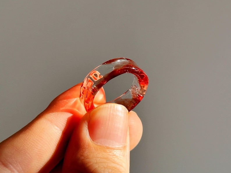 Pode incluir: Um anel de resina transparente com detalhes vermelhos e a palavra "ae" incorporada no design. O anel &eacute; exibido em um fundo cinza, destacando suas caracter&iacute;sticas transparentes e coloridas.