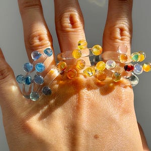 May include: A hand wearing multiple clear rings adorned with colorful, spherical beads. The beads are in shades of blue, yellow, orange, and red. The rings appear to be made of clear plastic or resin, and the overall effect is playful and artistic.