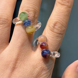 May include: Two clear glass rings, each adorned with colorful, spherical beads. One ring features green, blue, and yellow beads, while the other showcases peach, red, and blue beads. The rings are worn on fingers.
