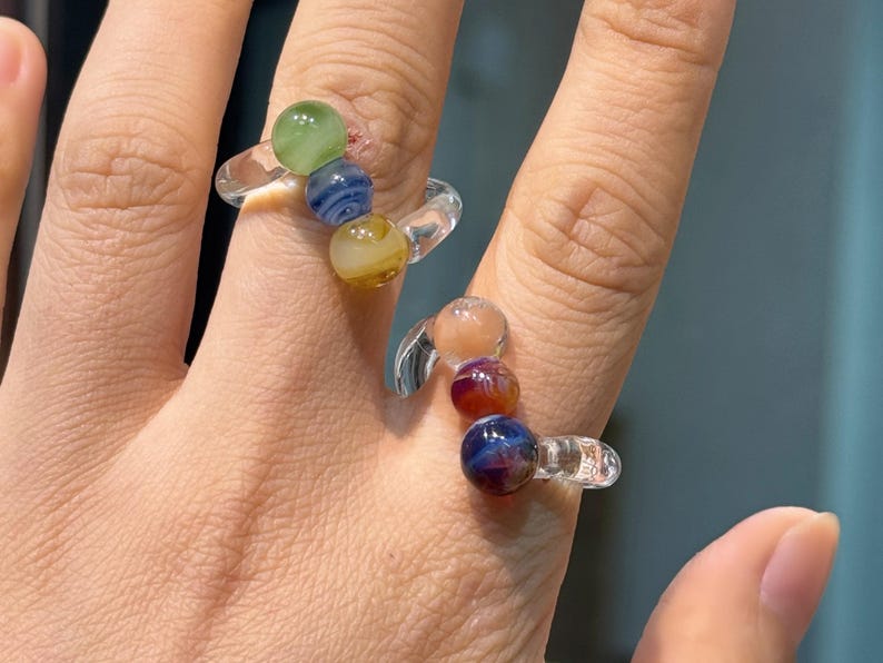 May include: Two clear glass rings, each adorned with colorful, spherical beads. One ring features green, blue, and yellow beads, while the other showcases peach, red, and blue beads. The rings are worn on fingers.