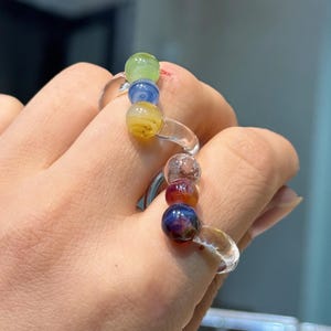 May include: Two glass bead rings are worn on a hand. One ring has green, blue, and yellow beads. The other ring has clear, red, and dark blue beads. The rings have clear bands.