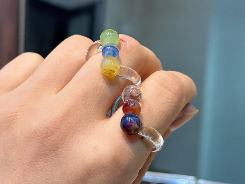 May include: Two glass bead rings are worn on a hand. One ring has green, blue, and yellow beads. The other ring has clear, red, and dark blue beads. The rings have clear bands.