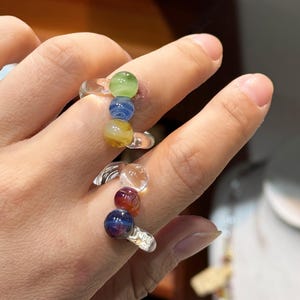 May include: Clear glass rings with colorful, spherical beads. One ring features green, blue, and yellow beads. The other ring has clear, red, and dark blue beads. The rings are worn on fingers.