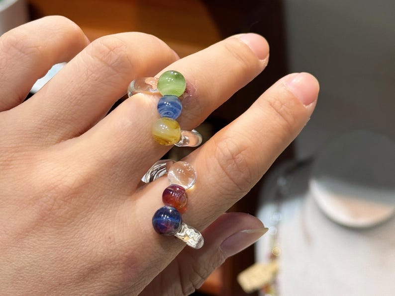 May include: Clear glass rings with colorful, spherical beads. One ring features green, blue, and yellow beads. The other ring has clear, red, and dark blue beads. The rings are worn on fingers.