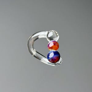 May include: Clear glass earring with a curved design. The earring features three colorful, spherical beads in shades of orange, red, and blue. The earring is displayed against a gray background.