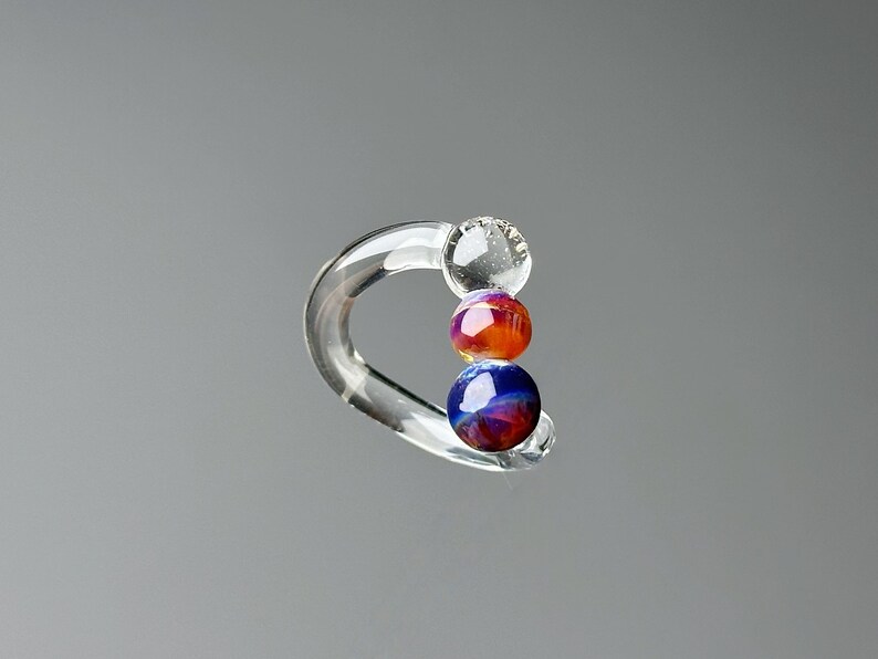 May include: Clear glass earring with a curved design. The earring features three colorful, spherical beads in shades of orange, red, and blue. The earring is displayed against a gray background.