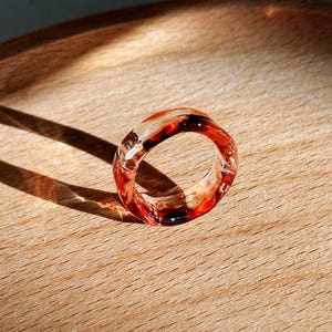 Pode incluir: Um anel de resina transparente com tons vermelhos e castanhos. O anel &eacute; circular e repousa sobre uma superf&iacute;cie de madeira, com uma sombra projetada para a esquerda. O design do anel apresenta uma mistura de cores. Joalharia.