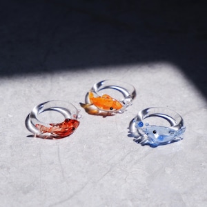 Peut inclure: Trois bagues en verre transparent, chacune avec un petit motif de poisson coloré à l'intérieur. Une bague présente un poisson rouge, une autre un poisson orange et la troisième un poisson bleu. Les bagues sont posées sur une surface gris clair.