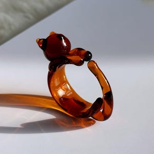May include: A unique amber-colored glass ring in the shape of a bear. The bear's head and paws are detailed, and the ring band curves around to form the bear's body. The ring is a decorative accessory.
