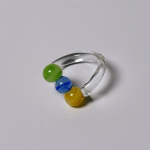 May include: A clear glass ring-shaped rattle with three colorful glass beads. The beads are green, blue, and yellow. The rattle is designed for infants and is a simple, yet visually appealing toy.