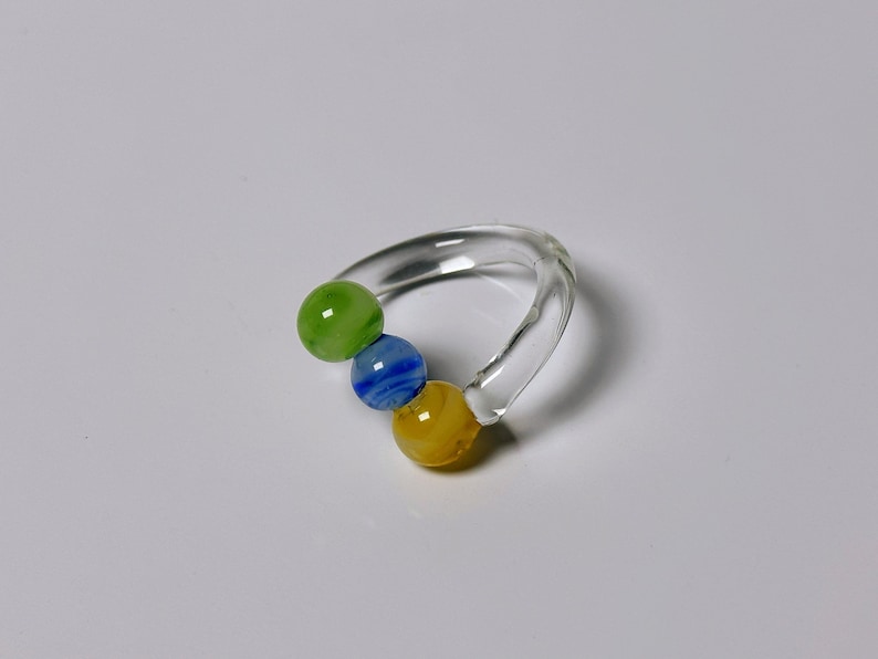 May include: A clear glass ring-shaped rattle with three colorful glass beads. The beads are green, blue, and yellow. The rattle is designed for infants and is a simple, yet visually appealing toy.