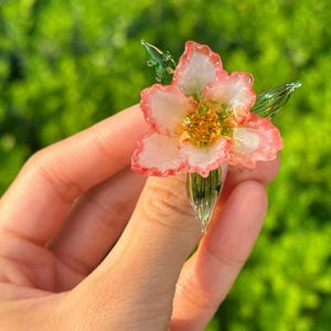 May include: A delicate glass flower with pink and white petals and a golden centre. The flower has green leaves and stem. The image is set against a blurred green background.