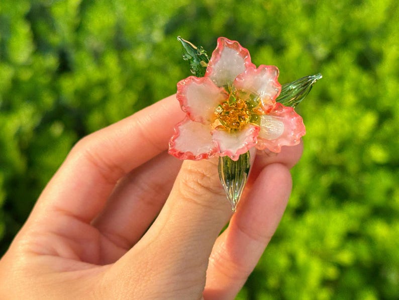 May include: A delicate glass flower with pink and white petals and a golden centre. The flower has green leaves and stem. The image is set against a blurred green background.
