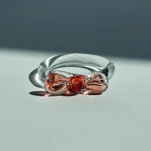 May include: A clear glass ring with a bow-shaped accent in shades of red and pink. The ring is transparent, allowing light to pass through, and sits on a white surface.
