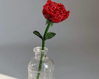 May include: A red glass rose with a green stem and leaves is displayed in a clear glass vase. The vase has an embossed floral design and sits on a small, round, woven coaster. The background is a plain white surface.