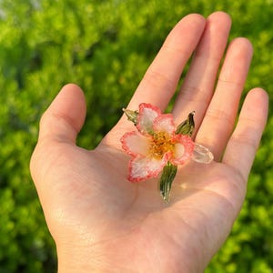 May include: A handcrafted glass flower with pink and white petals and a yellow centre. The flower has green glass leaves and is held in an open hand against a blurred green background. The flower is a unique piece of art.