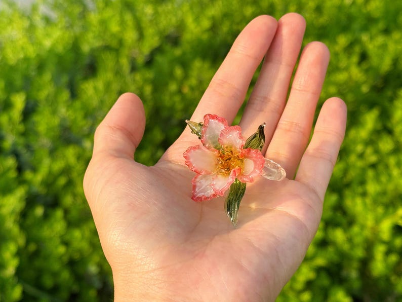 May include: A handcrafted glass flower with pink and white petals and a yellow centre. The flower has green glass leaves and is held in an open hand against a blurred green background. The flower is a unique piece of art.