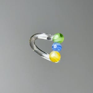 May include: A clear glass ring with three colorful glass beads. The beads are green, blue, and yellow. The ring is curved and has a smooth, polished finish. The beads are round and have a swirled pattern.