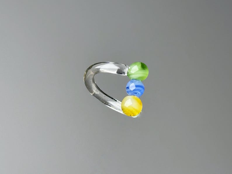 May include: A clear glass ring with three colorful glass beads. The beads are green, blue, and yellow. The ring is curved and has a smooth, polished finish. The beads are round and have a swirled pattern.