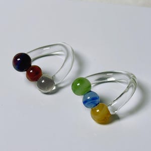May include: Two clear glass napkin rings, each with three colorful glass beads. One ring has a dark blue, red, and clear bead. The other has green, blue, and yellow beads. The rings are on a white surface.