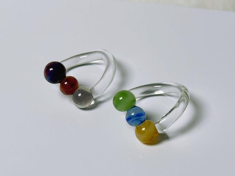 May include: Two clear glass napkin rings, each with three colorful glass beads. One ring has a dark blue, red, and clear bead. The other has green, blue, and yellow beads. The rings are on a white surface.