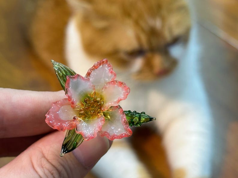 May include: A handcrafted glass flower with white petals edged in pink, a yellow centre, and green leaves. The delicate floral design is held in a person's hand, with a blurred orange and white cat in the background.