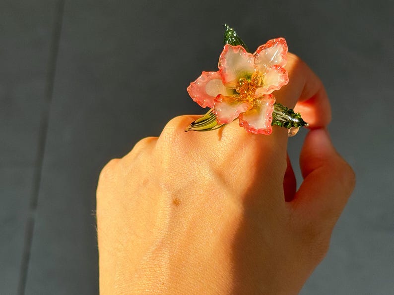 May include: A floral ring with a flower design. The ring has pink and white petals, with gold accents. Green leaves and stems complete the natural look. A unique piece of jewellery.
