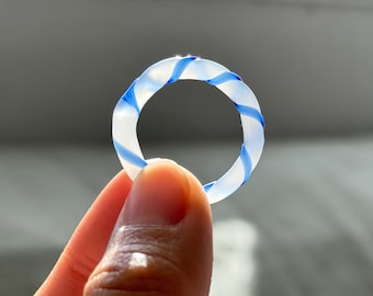May include: A clear ring with a blue and white spiral design. The ring is held up against a blurred background. The ring is a circular shape with a twisted pattern of blue and white.