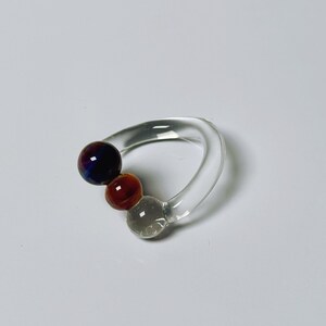 May include: A clear glass ring with three colorful, spherical accents. The largest sphere is a deep purple with blue and red hues, followed by a reddish-brown sphere, and a gray sphere. The ring has a curved, smooth design.