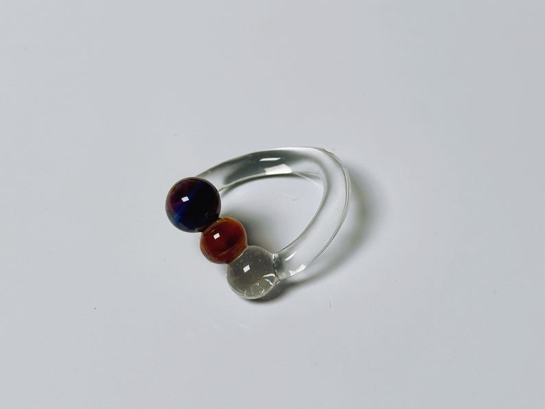 May include: A clear glass ring with three colorful, spherical accents. The largest sphere is a deep purple with blue and red hues, followed by a reddish-brown sphere, and a gray sphere. The ring has a curved, smooth design.