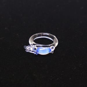 May include: A clear glass ring featuring a small, sculpted blue and white squid or shrimp figure attached to the band. The creature has a smooth, rounded body with a light blue center, a dark eye, and translucent tentacles extending forward.