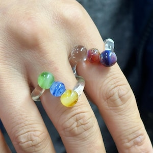 May include: Two clear glass rings, each adorned with colorful, spherical beads. One ring features green, blue, and yellow beads. The other ring has brown, red, and purple beads. The rings are worn on fingers.