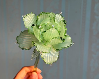 May include: A delicate, light green glass flower with ruffled edges and sparkling accents. The petals are translucent, revealing intricate details. The stem is a matching green. A decorative floral piece.