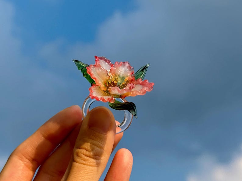 May include: A clear glass ring with a flower design. The flower has pink and yellow petals and green leaves. The ring is held against a blue sky. The ring is a piece of jewellery.