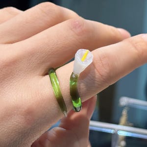 May include: A unique ring featuring a calla lily design. The ring has a green stem and a white flower with a yellow center. The ring is worn on a finger, showcasing the intricate details of the floral jewelry.