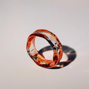 Pode incluir: Um anel de resina transparente com um design org&acirc;nico e rodopiante. O anel apresenta tons de laranja e vermelho, criando um efeito marmorizado. O anel est&aacute; sobre um fundo branco, projetando uma sombra.