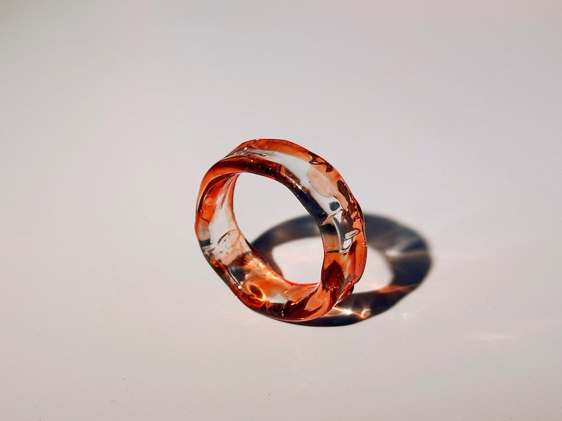 Pode incluir: Um anel de resina transparente com um design org&acirc;nico e rodopiante. O anel apresenta tons de laranja e vermelho, criando um efeito marmorizado. O anel est&aacute; sobre um fundo branco, projetando uma sombra.