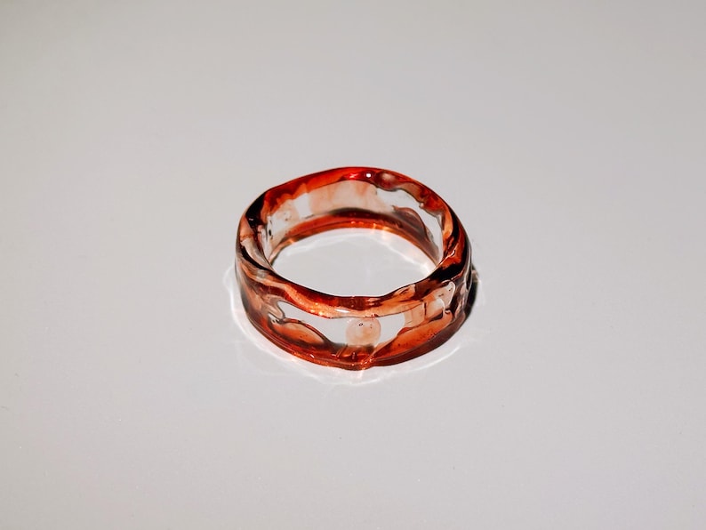 Pode incluir: Um anel transparente com um design &uacute;nico. O anel apresenta uma banda com uma apar&ecirc;ncia ondulada e texturizada, e &eacute; acentuado com tons de vermelho e laranja. O anel &eacute; uma joia.